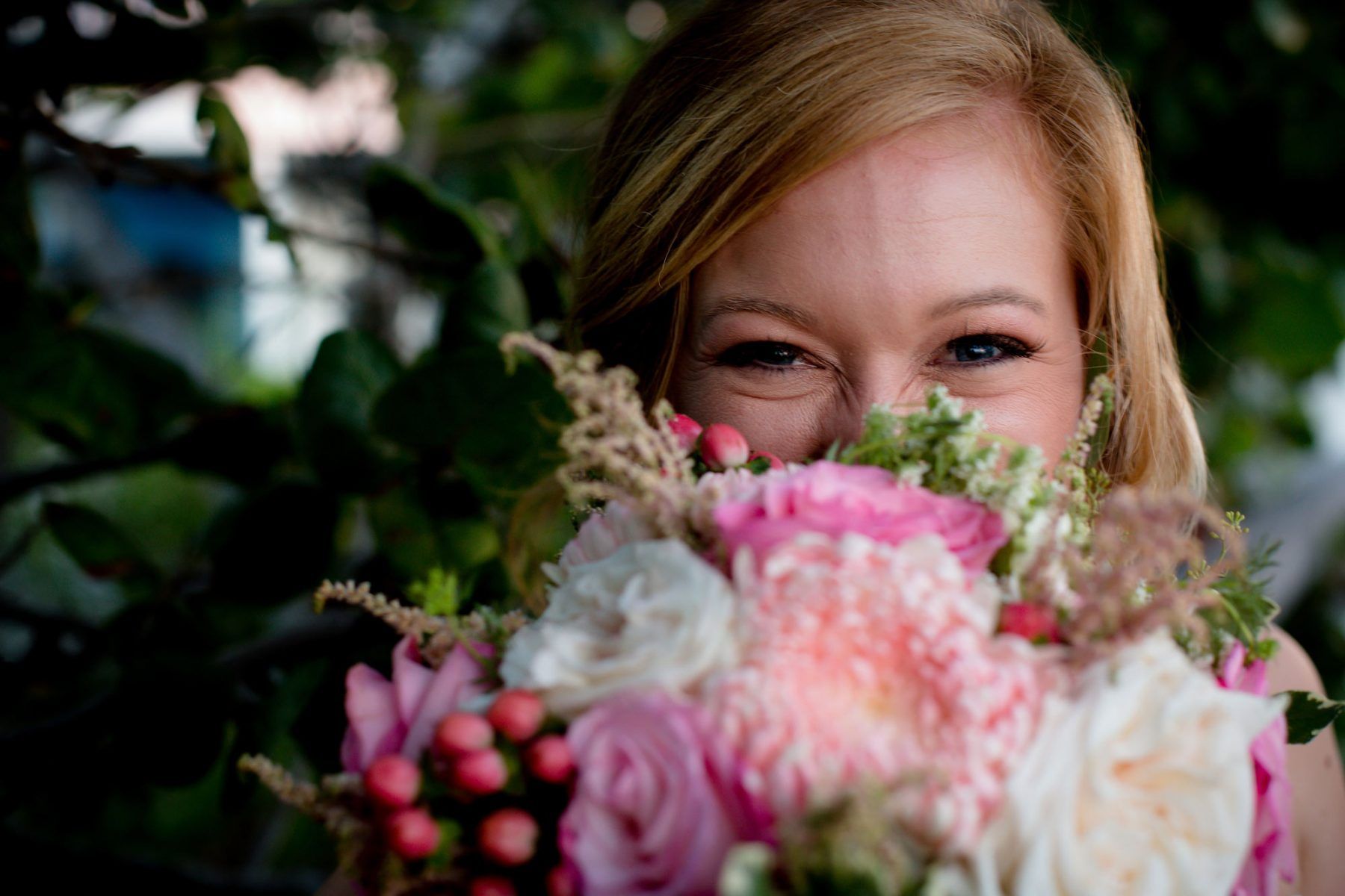 Woman peeking over a bouquet of pink and white flowers; blurred greenery in background.