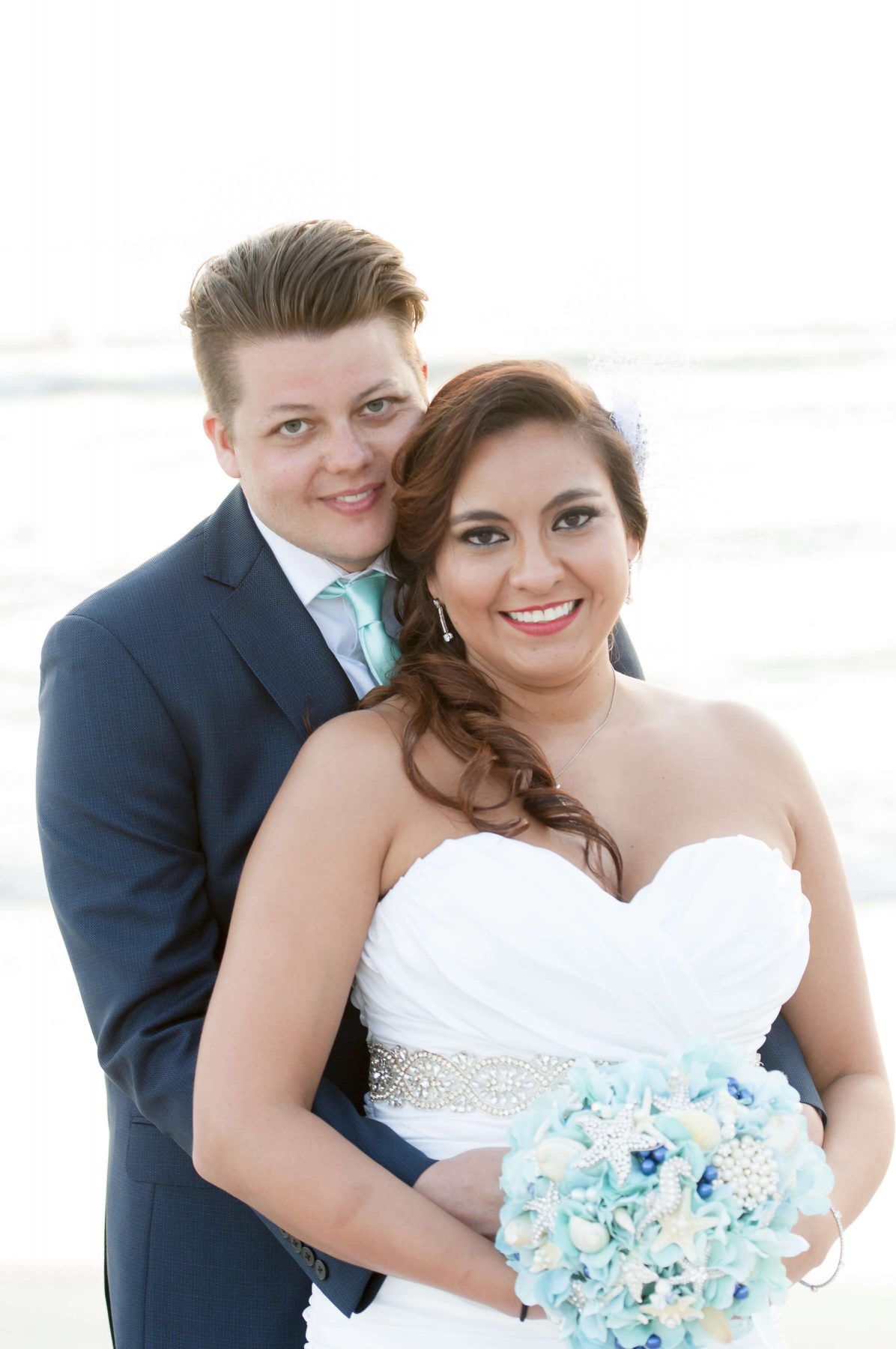 Bride and groom embrace on beach; bride wears white strapless dress, groom in blue suit.