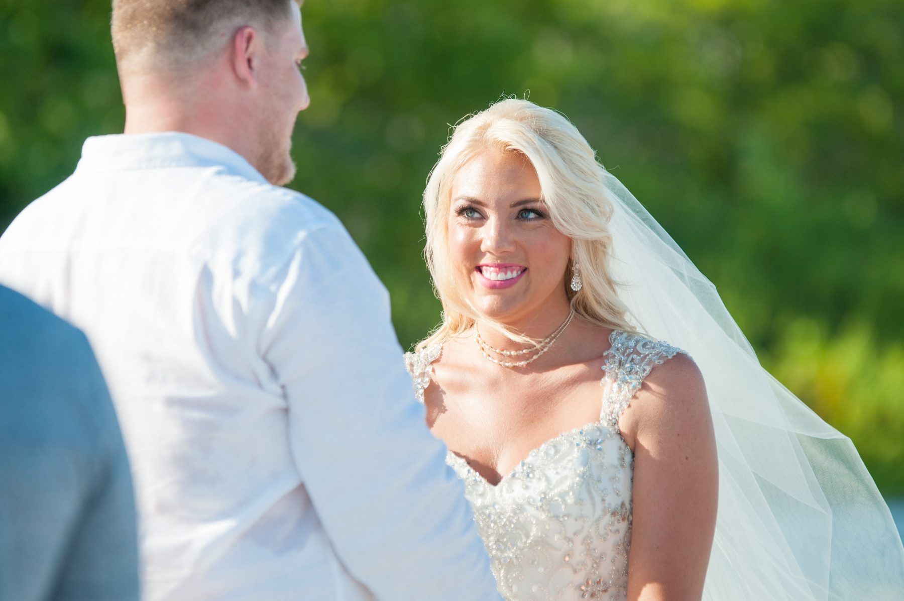 Bride smiles at groom during a beach wedding. She wears a beaded dress and veil, he wears white shirt.