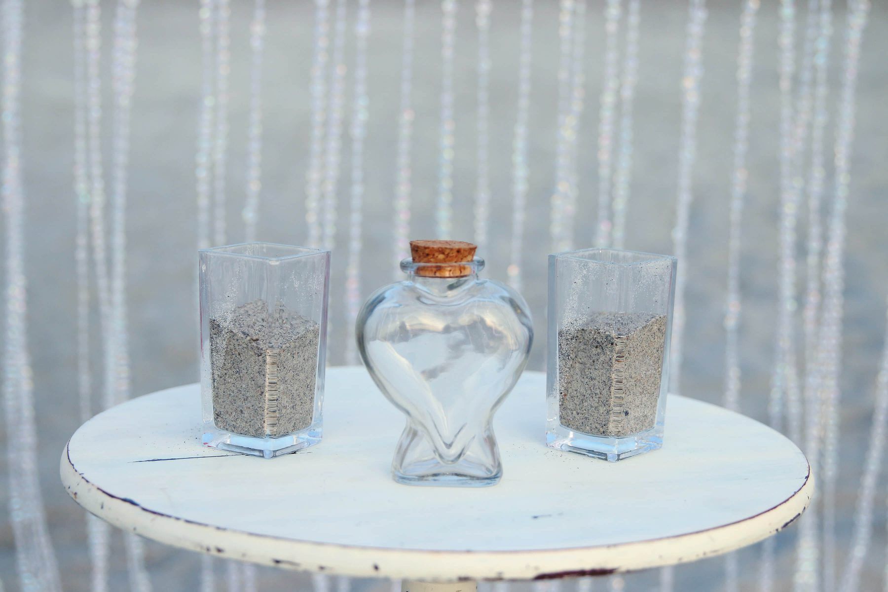 Wedding sand ceremony setup: two glasses of grey sand, heart-shaped container with white sand, on a white table.