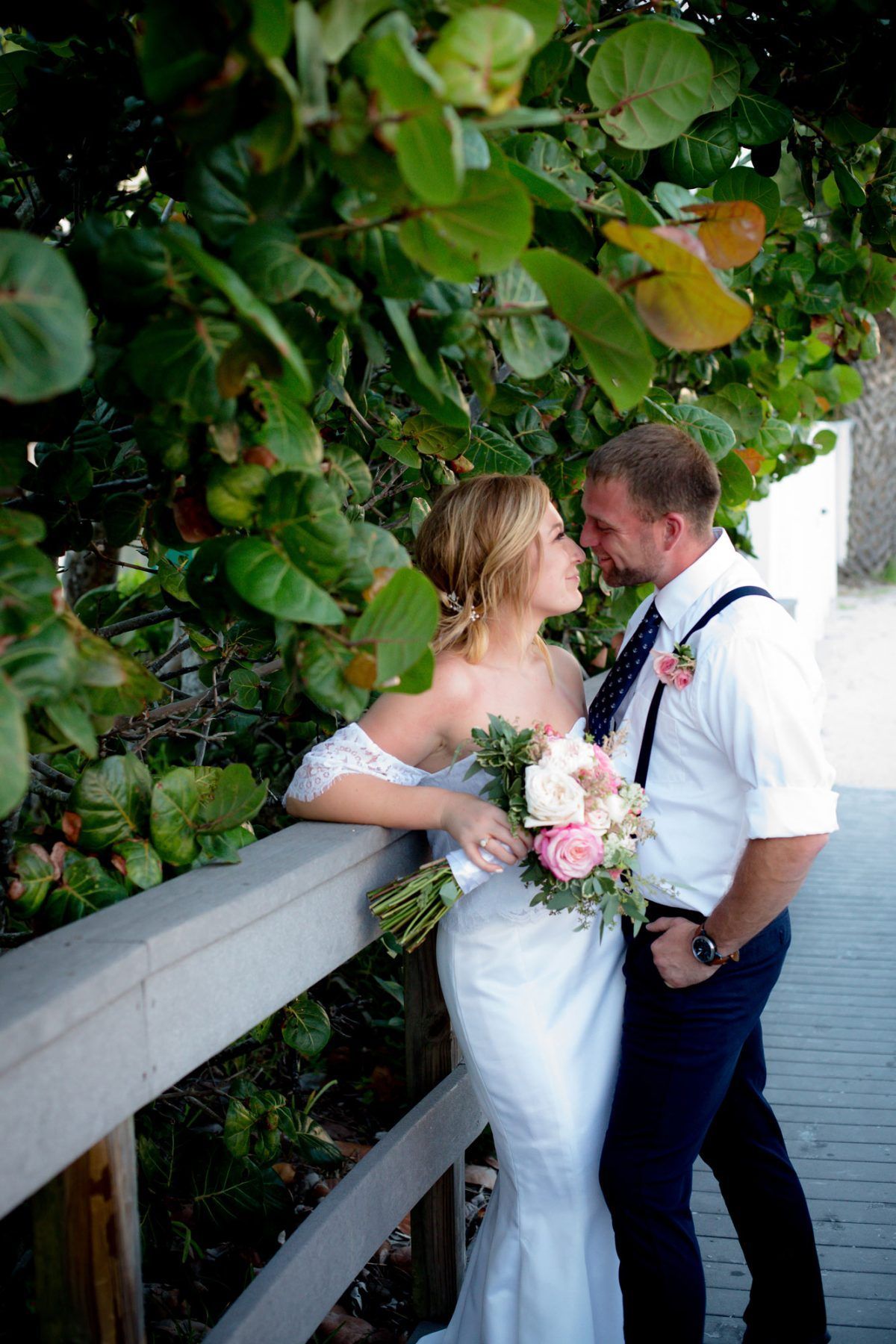 Bride and groom kissing near a wooden fence with greenery overhead. The bride holds flowers and wears a white dress.