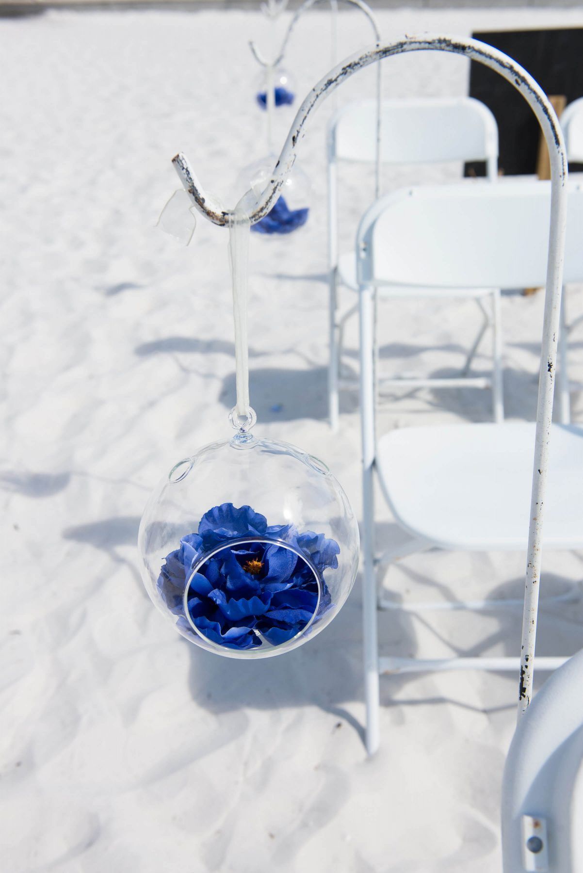 Clear glass orb with blue petals hanging on a white chair at a beach wedding.