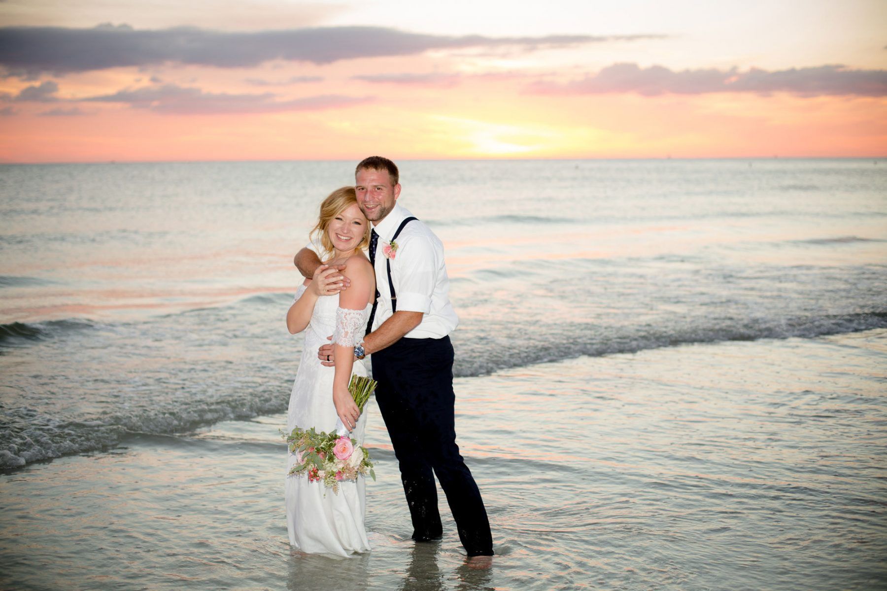 Couple on a beach embracing at sunset. Woman in a white dress, man in suspenders. Waves and colorful sky.