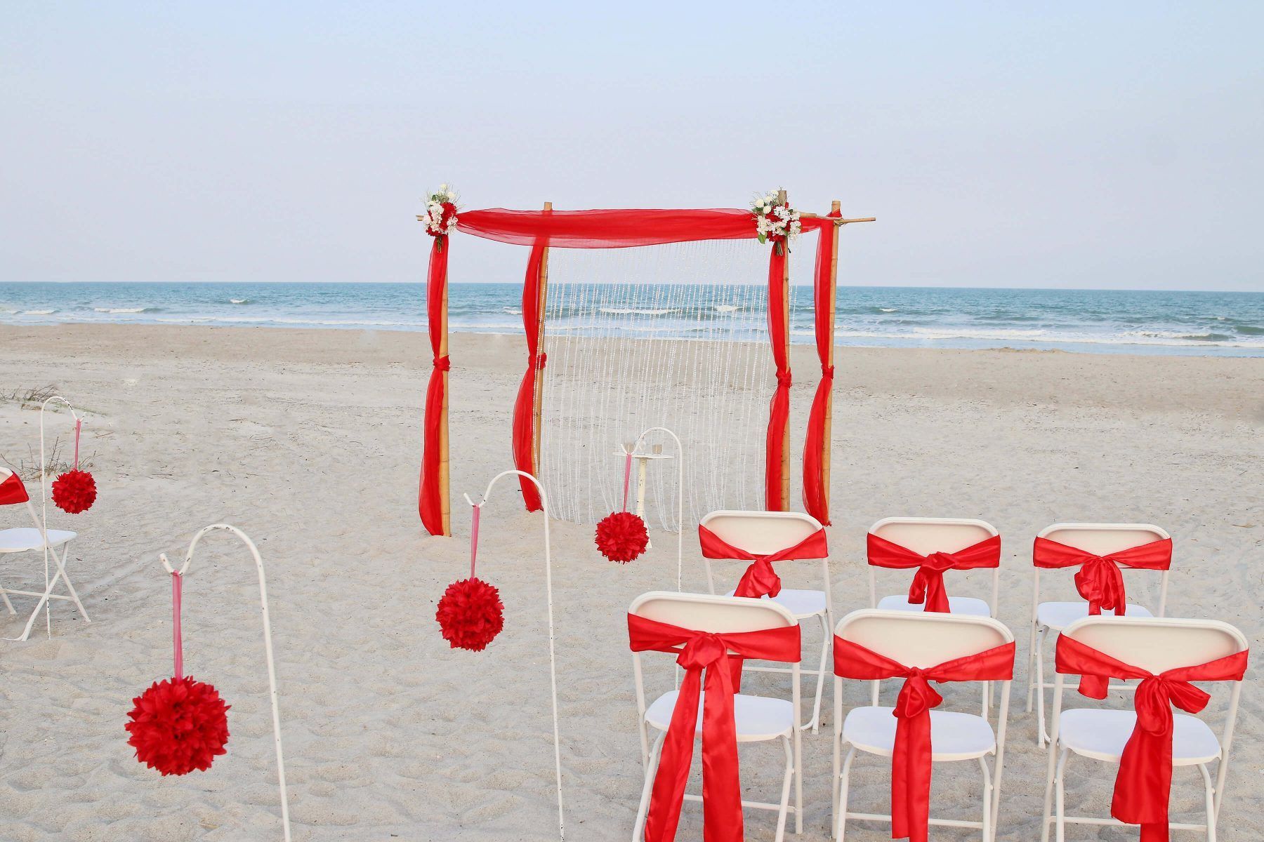 Beach wedding setup with red draping, chairs, and decorations. Ocean in background.