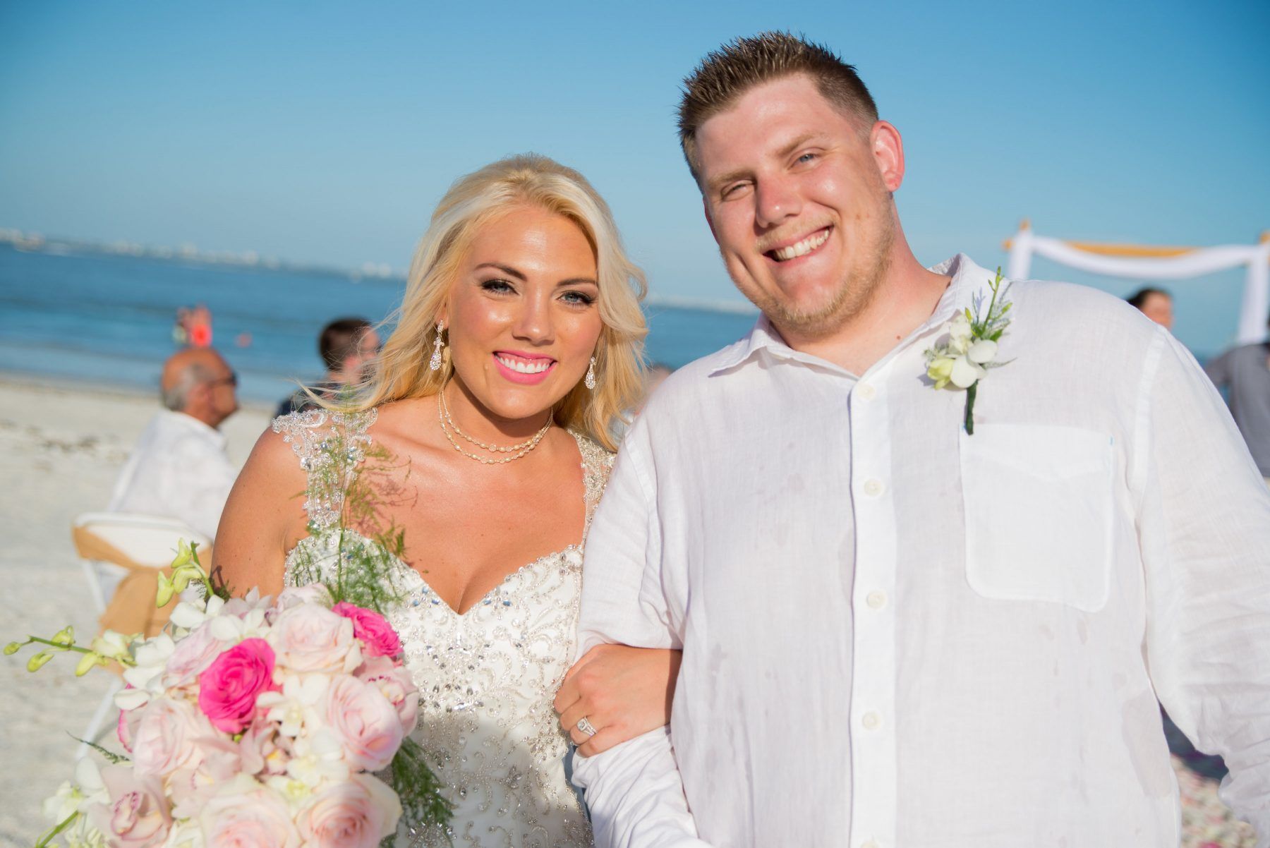 Bride and groom smiling on a beach; she wears a wedding dress, he wears a white shirt.