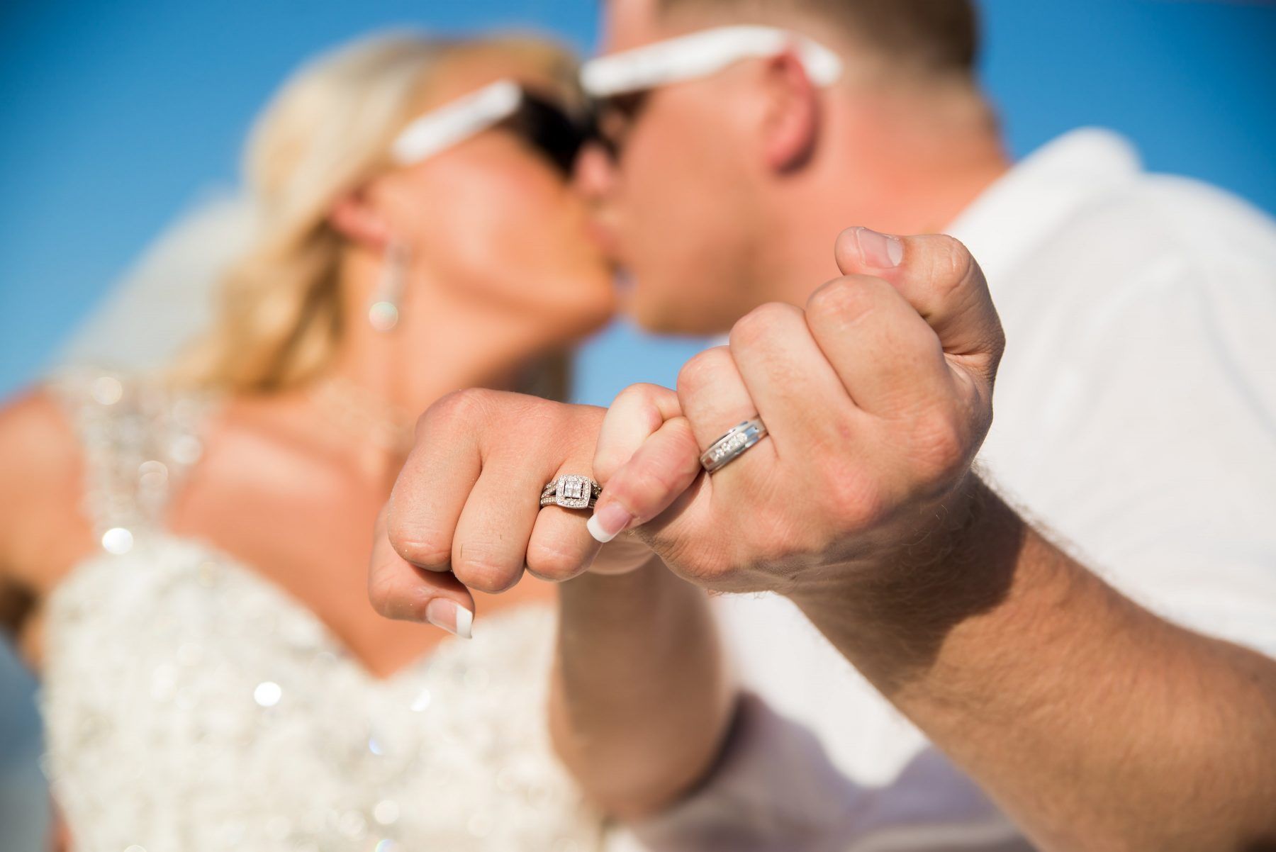 Couple kissing, showing wedding rings on their fingers; clear blue sky background.