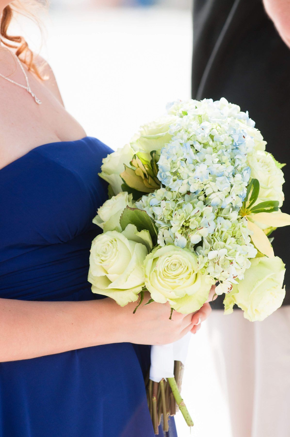 Woman in a blue dress holding a bouquet of white roses and blue hydrangeas, outdoor setting.