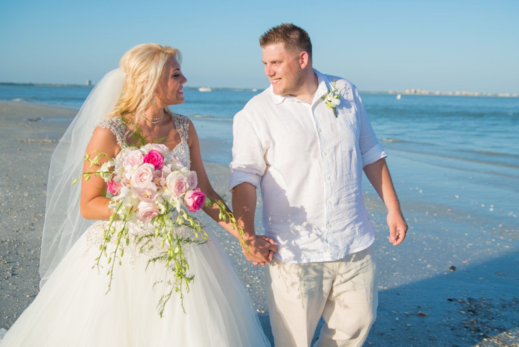 Bride and groom holding hands, smiling, walking on a beach. She wears a white dress and veil; he wears a white shirt.