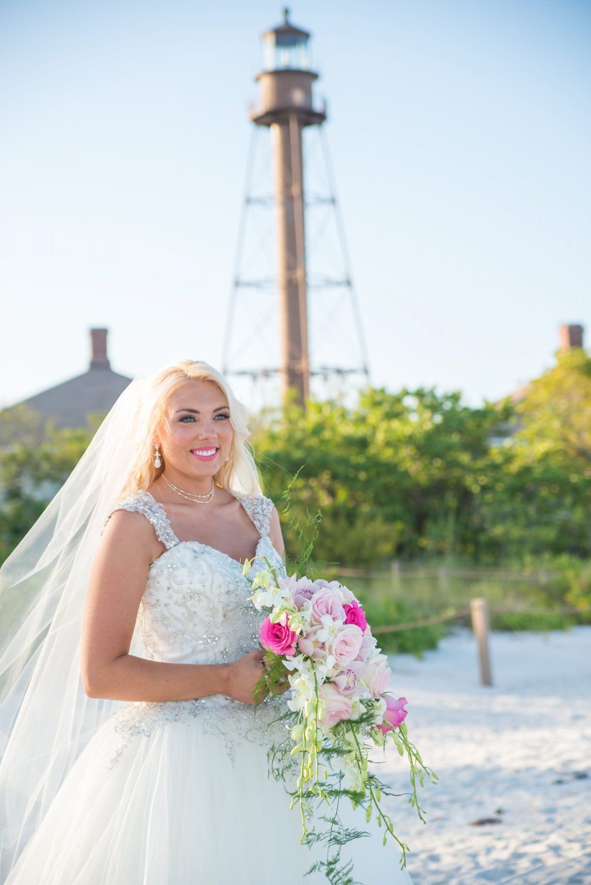 Bride in a white gown holds bouquet, smiling on a beach with a lighthouse in the background.