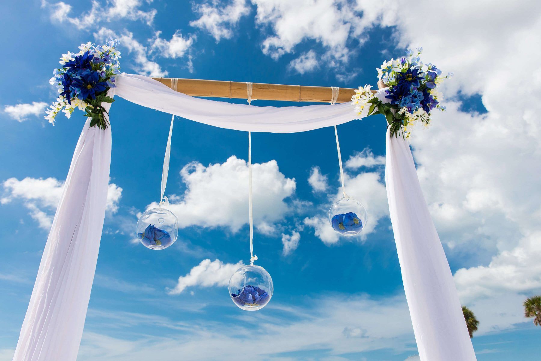 Wedding arch with blue flowers and hanging glass globes against a blue sky with clouds.