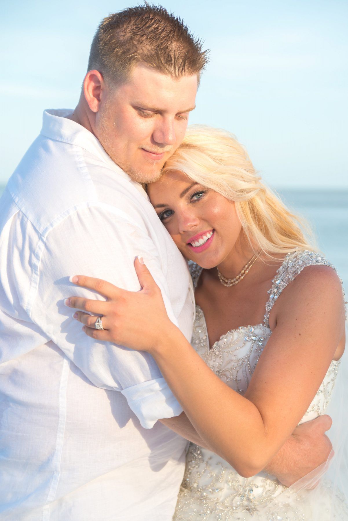 Couple embracing on a beach; woman in wedding dress, man in white shirt.
