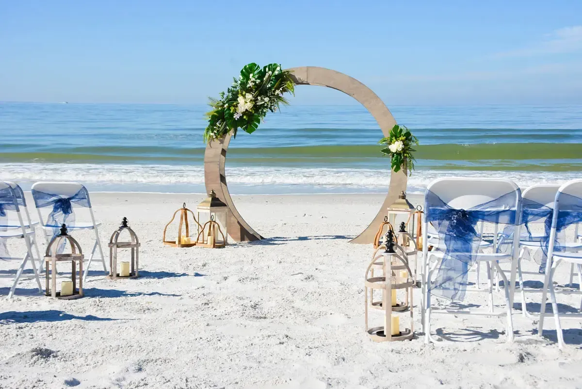 Beach wedding setup with a floral arch, chairs, and lanterns on the sand, with ocean in the background.
