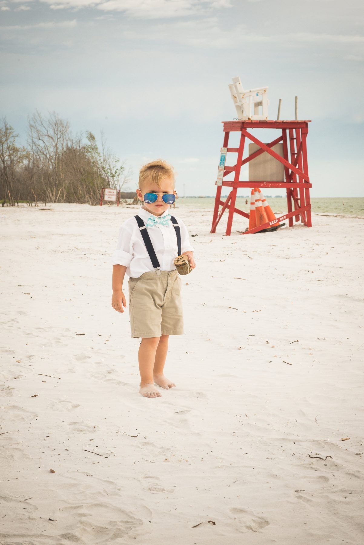 Young child in sunglasses, suspenders, and shorts stands on a sandy beach near a red lifeguard stand.