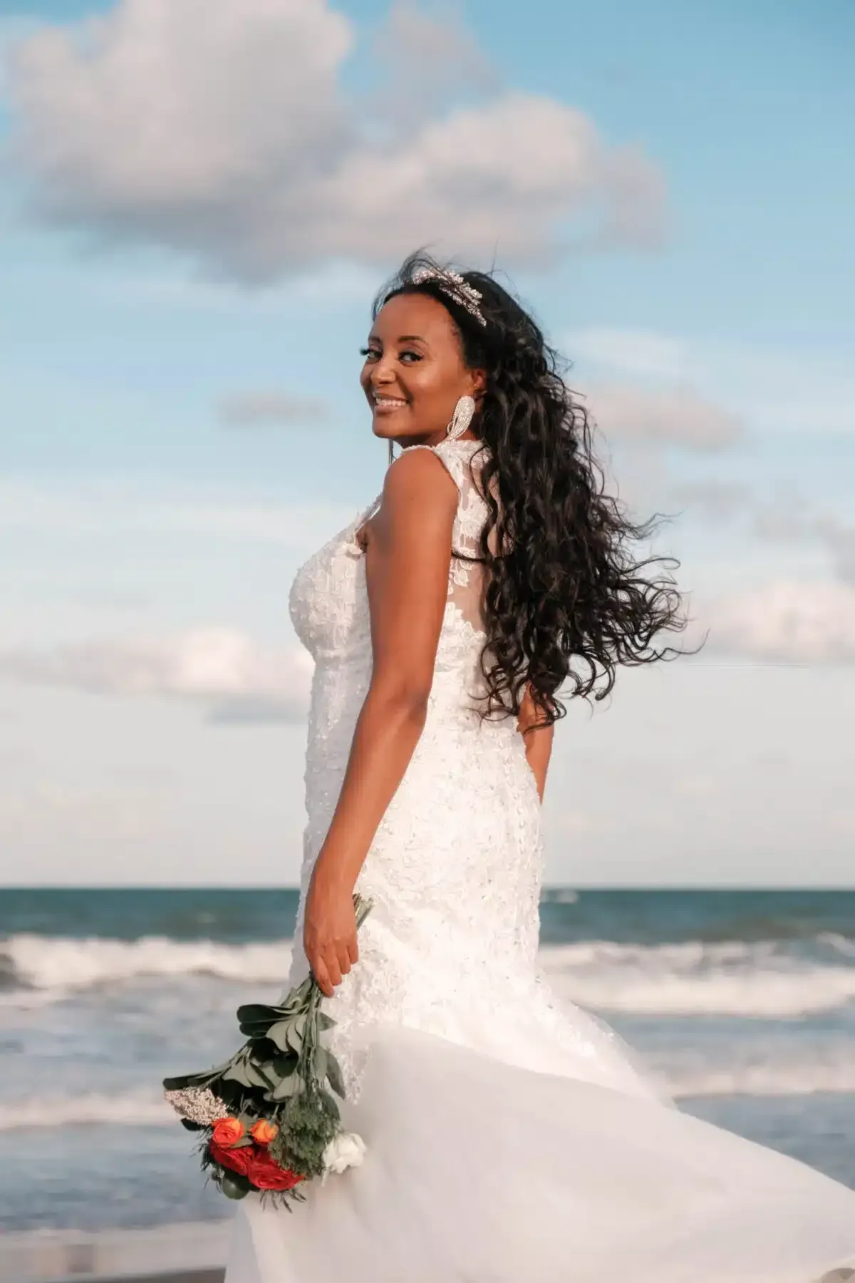 Bride in a white gown smiles at the camera on a beach; holding flowers.