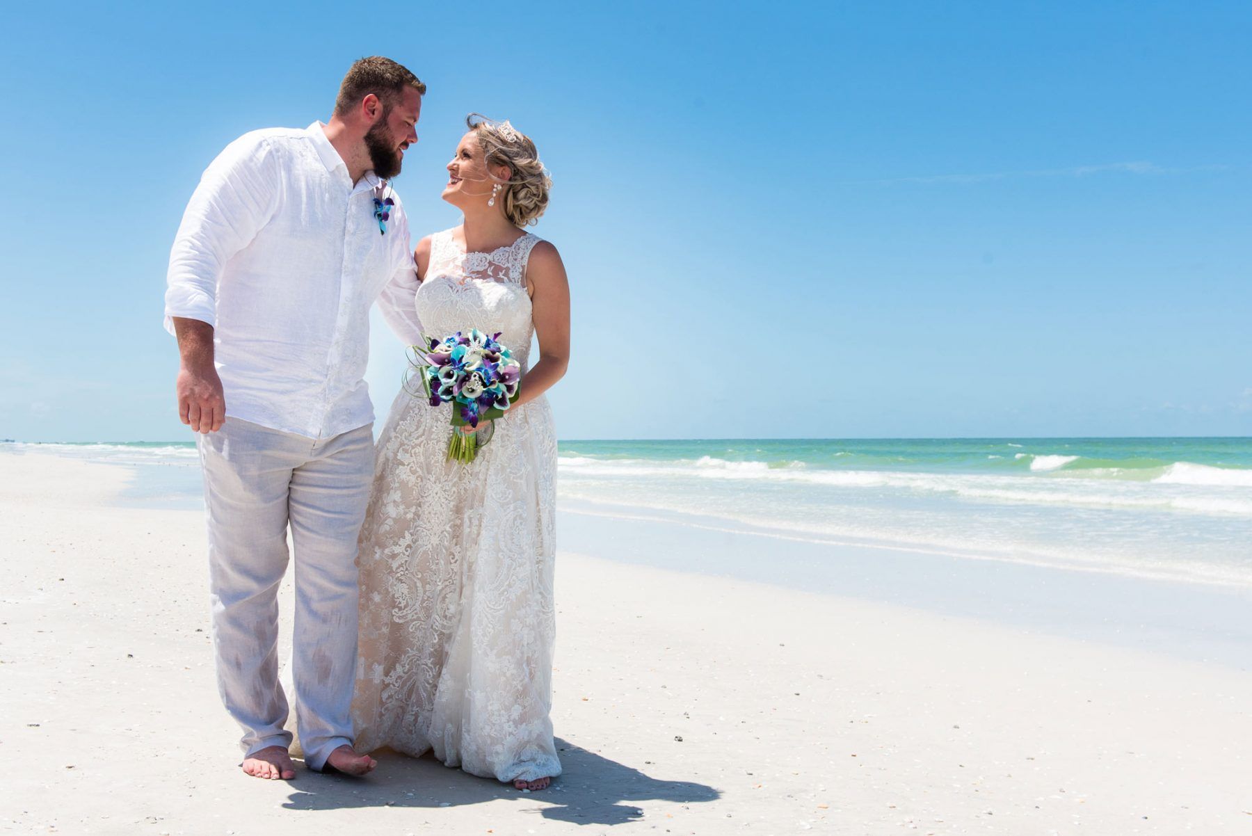 Couple in wedding attire on a beach, gazing at each other. Clear blue sky and ocean in the background.