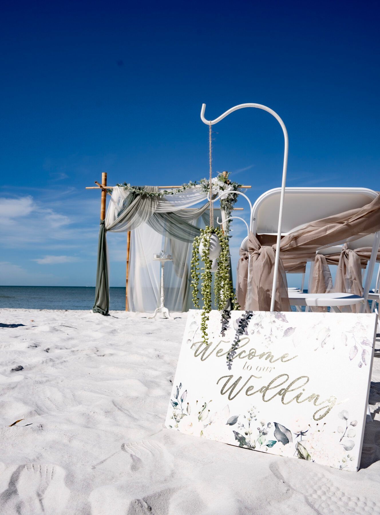 Beach wedding setup with welcome sign and decorated chairs, against a blue sky.