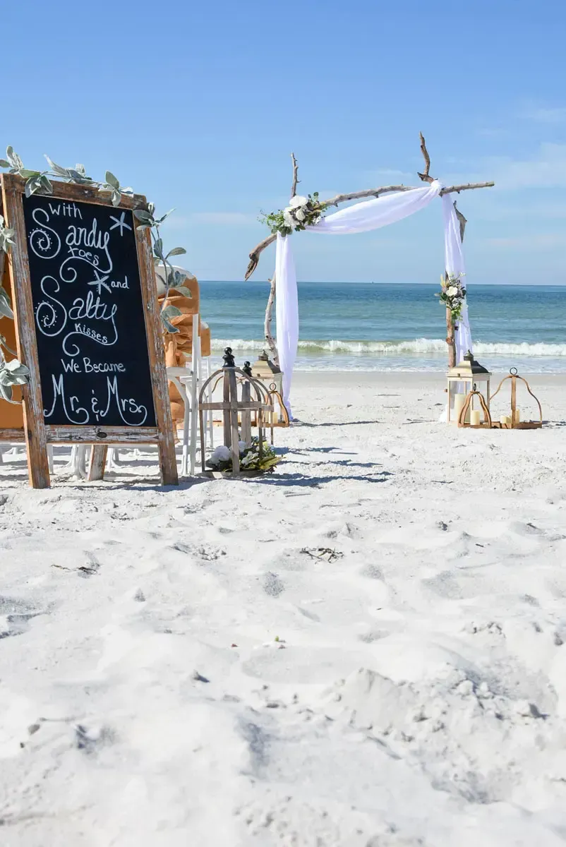 Beach wedding setup with chalkboard sign, floral arch, and white sand, with ocean in background.