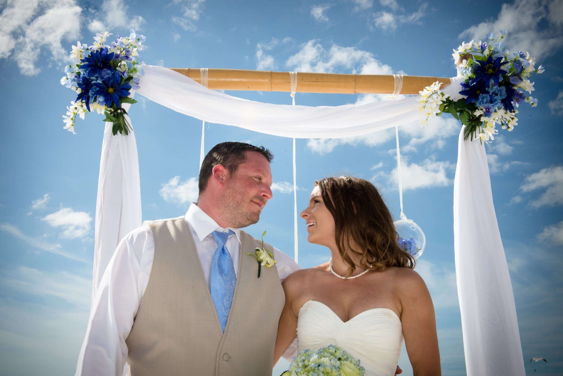 Couple at a beach wedding, looking at each other under an arch decorated with flowers and fabric, blue sky backdrop.