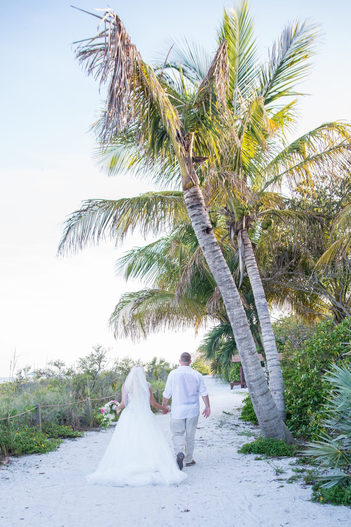 Bride and groom walk hand-in-hand on a sandy path, past palm trees, towards the horizon.