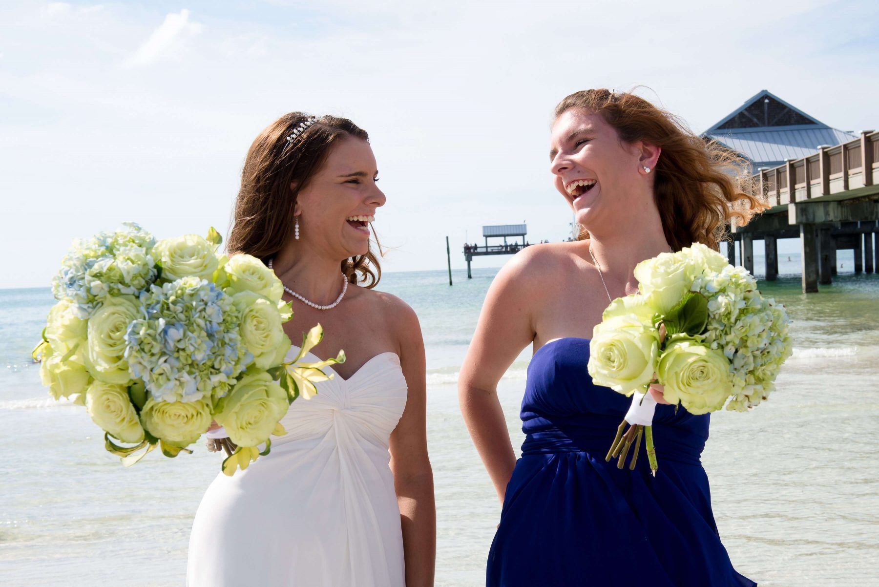 Bride and bridesmaid laughing on beach, holding bouquets. Bride in white, bridesmaid in blue. Pier in background.