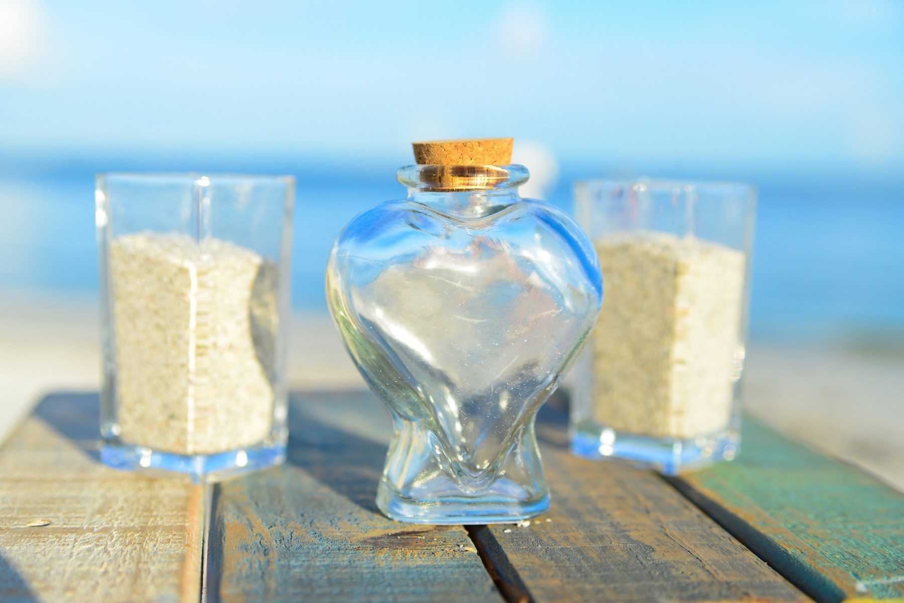 Heart-shaped glass bottle with a cork on a wooden surface, flanked by two glasses of sand; beach in the background.