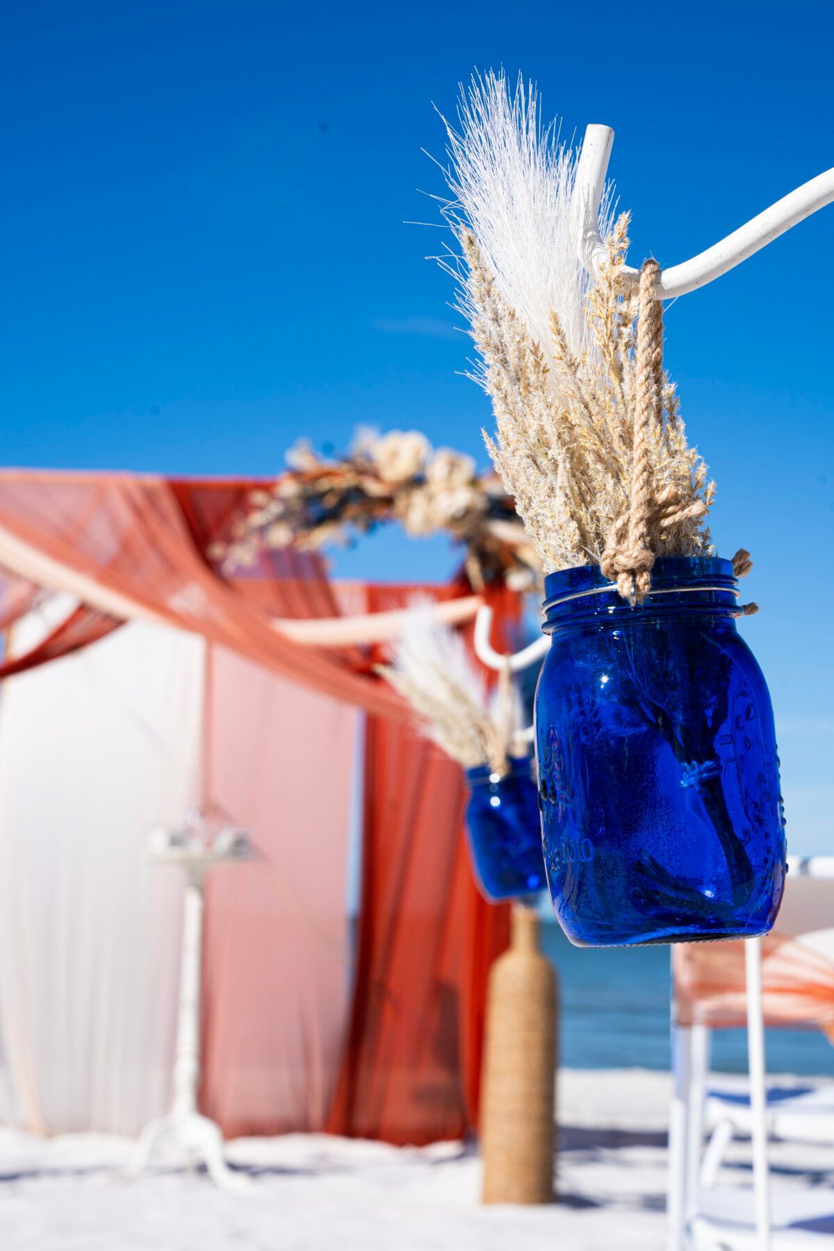 Blue mason jars with dried flowers hanging on a white branch, beach setting with blue sky.