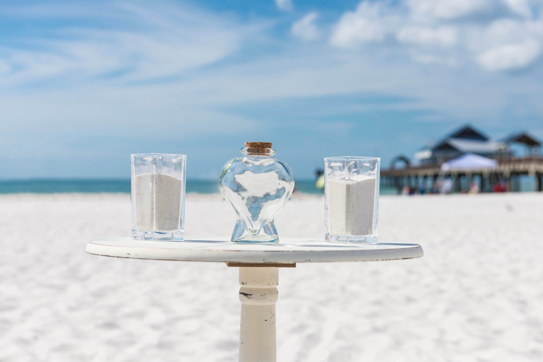 Sand ceremony on a beach; two glasses of sand and a central vessel, with ocean and blue sky in background.