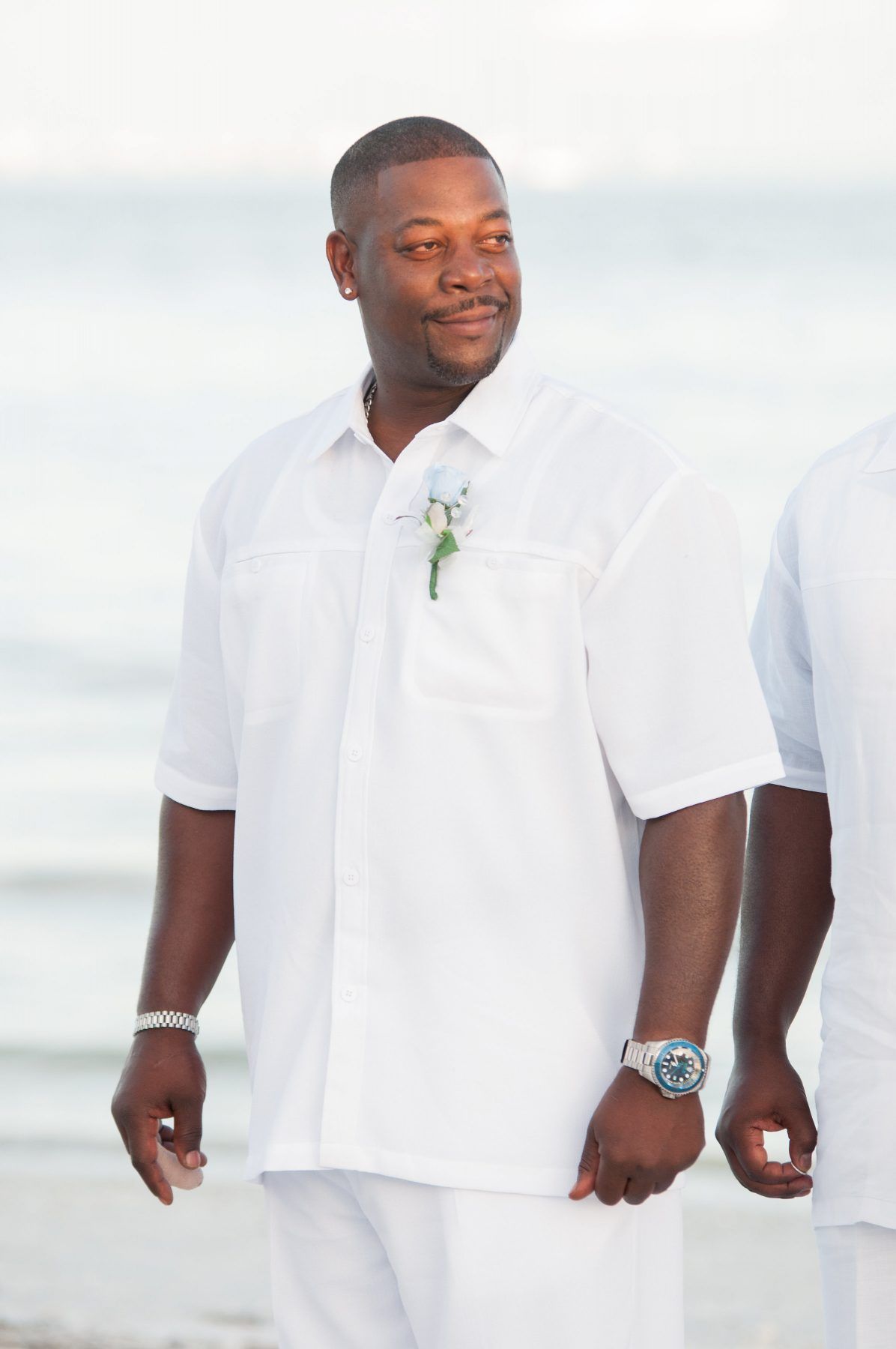 Man in white shirt and pants, looking to the side, wearing watch, flower on shirt, holding hands, beach background.