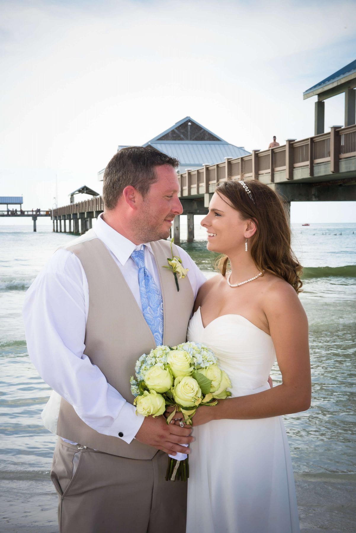 Bride and groom smiling at each other on a beach. She holds flowers, he wears a vest, a pier is in the background.