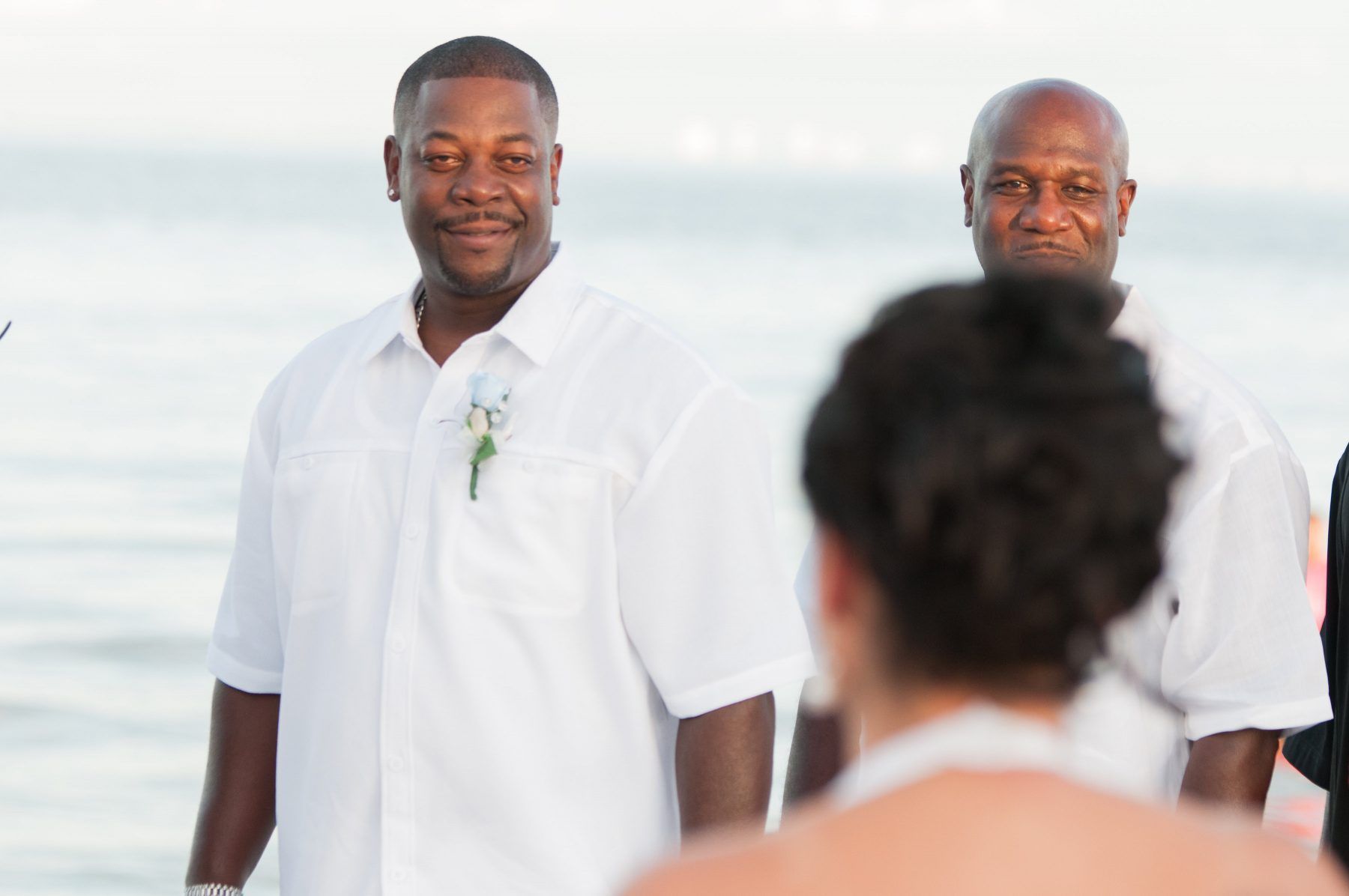Two men in white shirts, one smiling, at a beach ceremony.