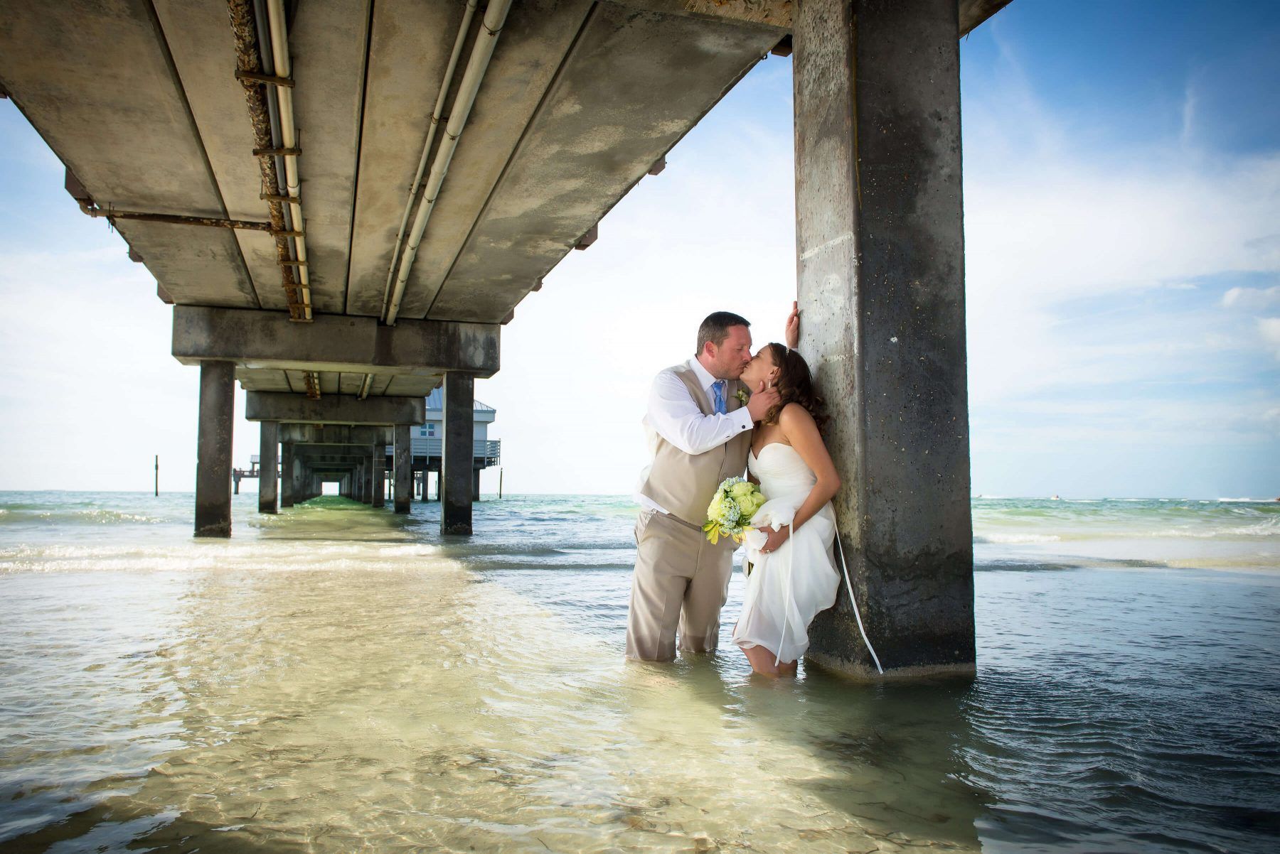 Couple in wedding attire under pier, standing in shallow water, man kissing woman's forehead, sunny beach.