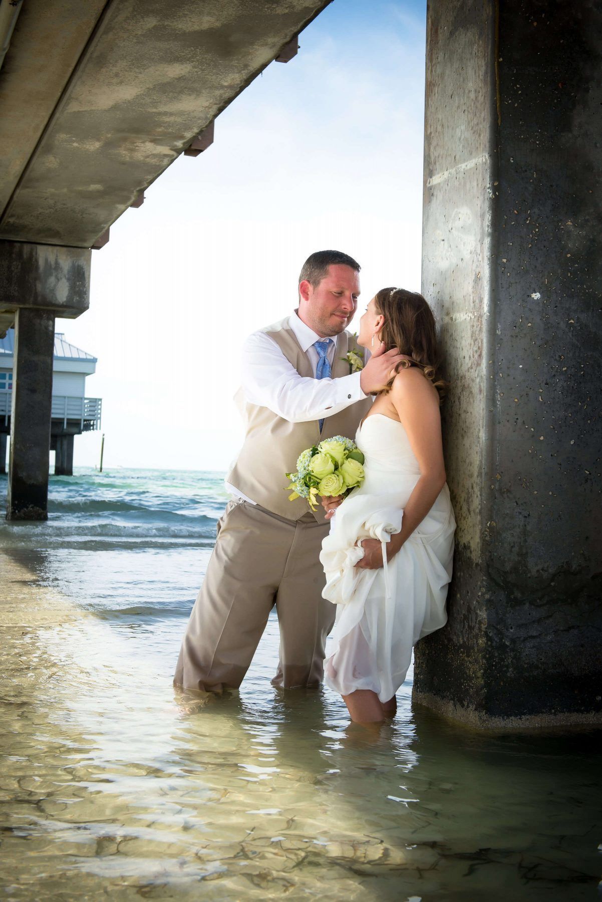 Groom and bride stand in shallow water under a pier, embracing. The bride holds a bouquet, the sky is bright.