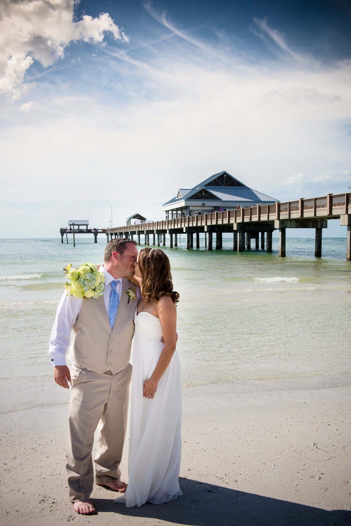 Bride and groom on beach, facing each other, with pier in background under blue sky.