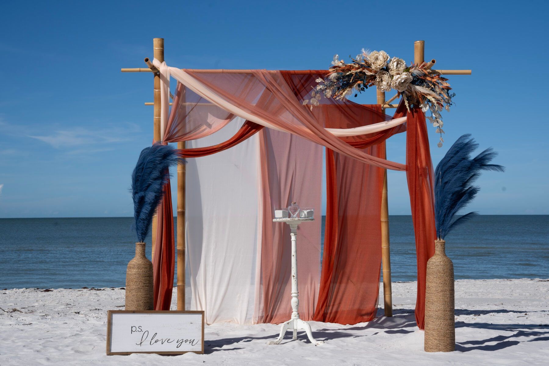 Beach wedding arch with rust-colored fabric, navy plumes, and floral arrangement against a blue sky.