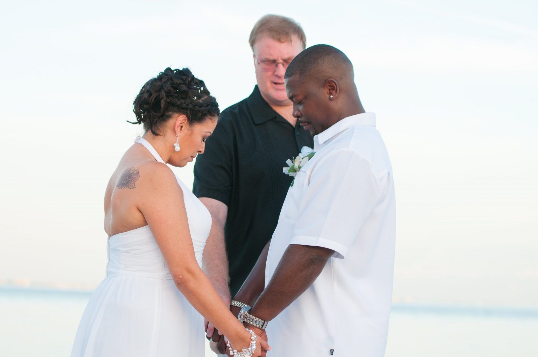 Couple in white, holding hands during a beach wedding ceremony, facing an officiant.