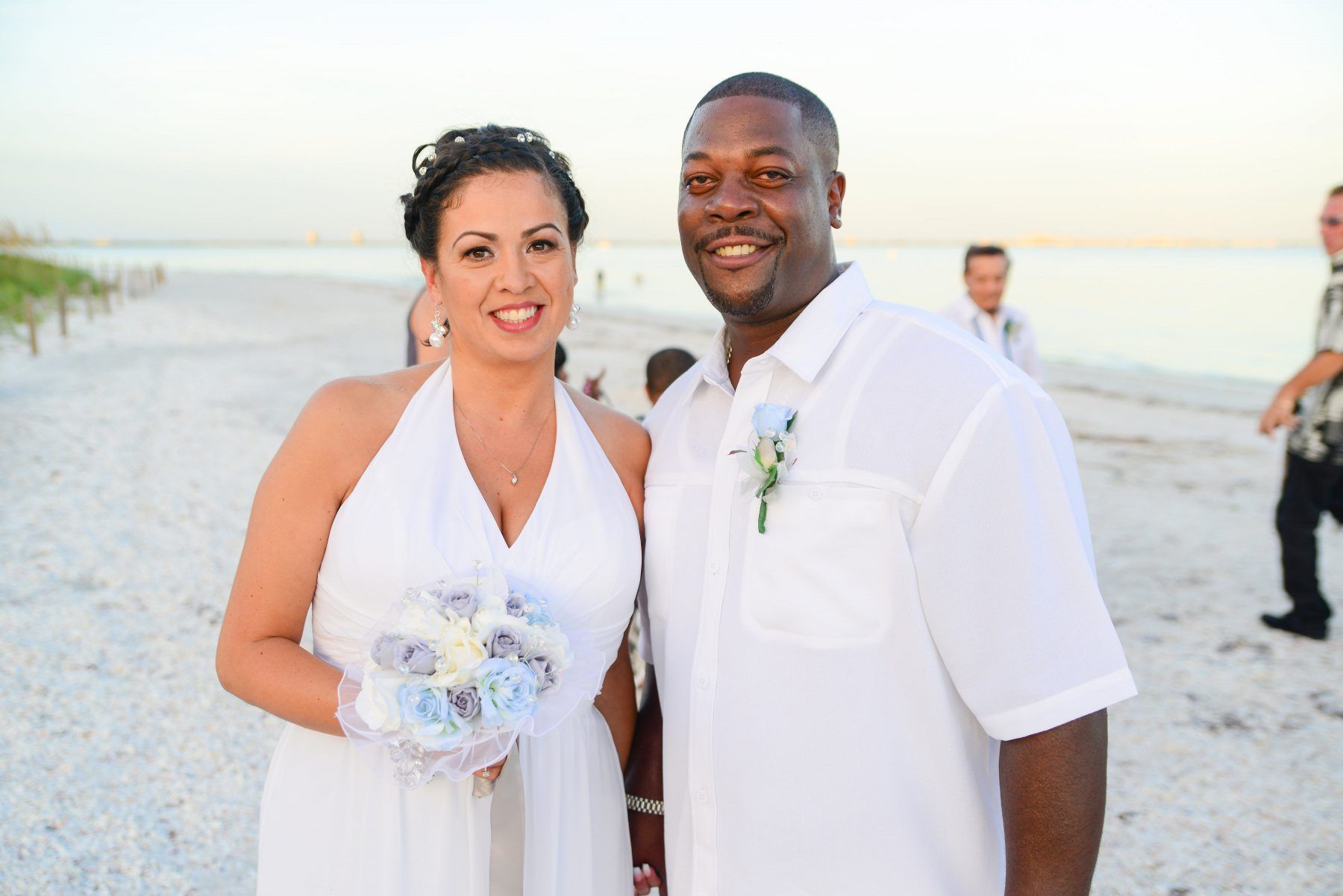 Couple at a beach wedding, bride holding bouquet, groom wearing a white shirt, smiling.