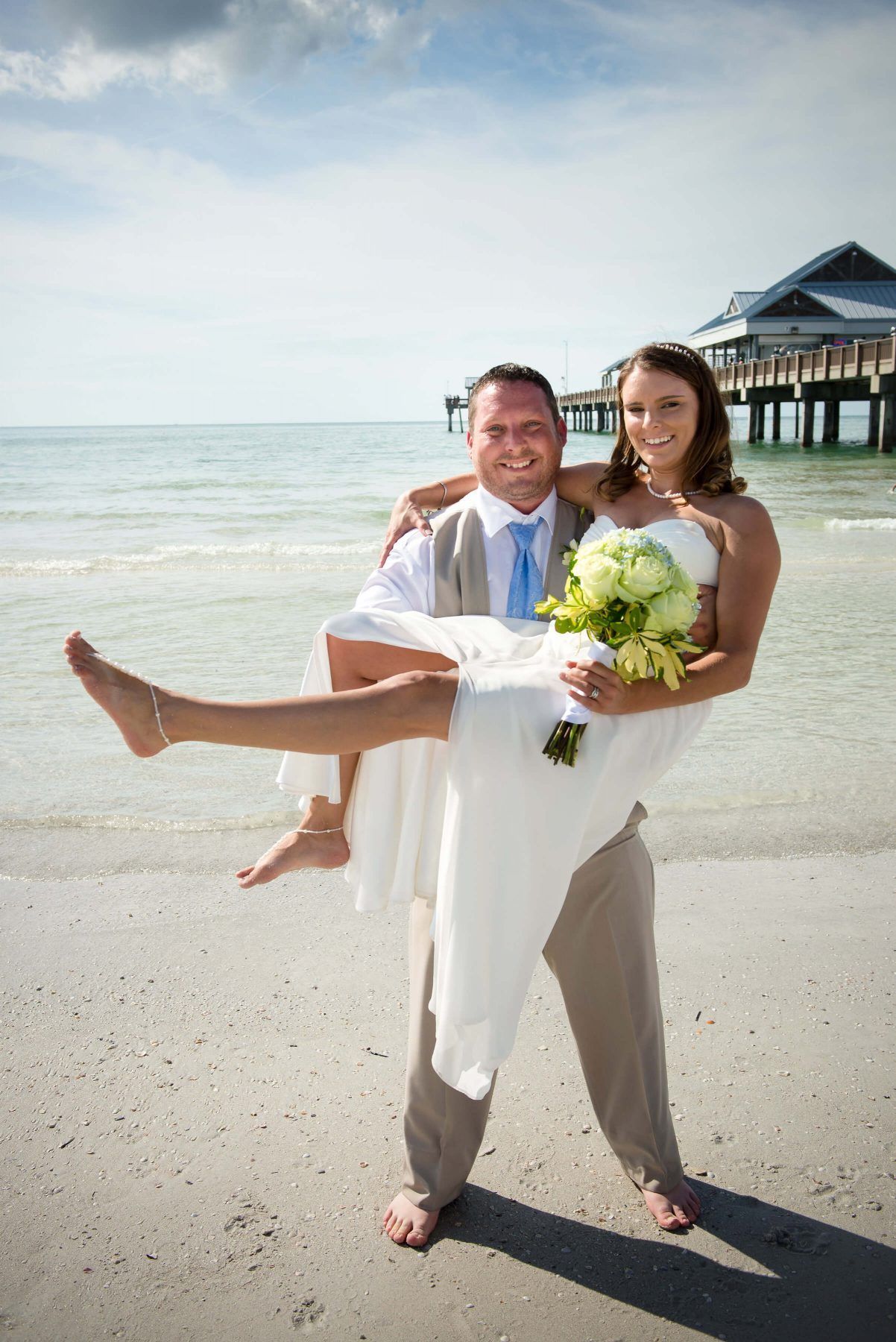 Groom carrying bride on beach; she holds flowers. Clear water, sunny sky, pier in background.