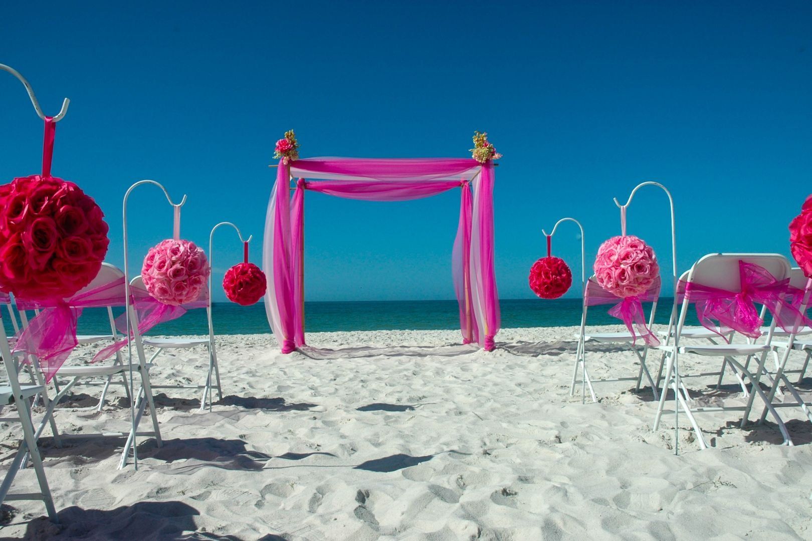 Beach wedding setup with pink decorations and arch against a clear blue sky.