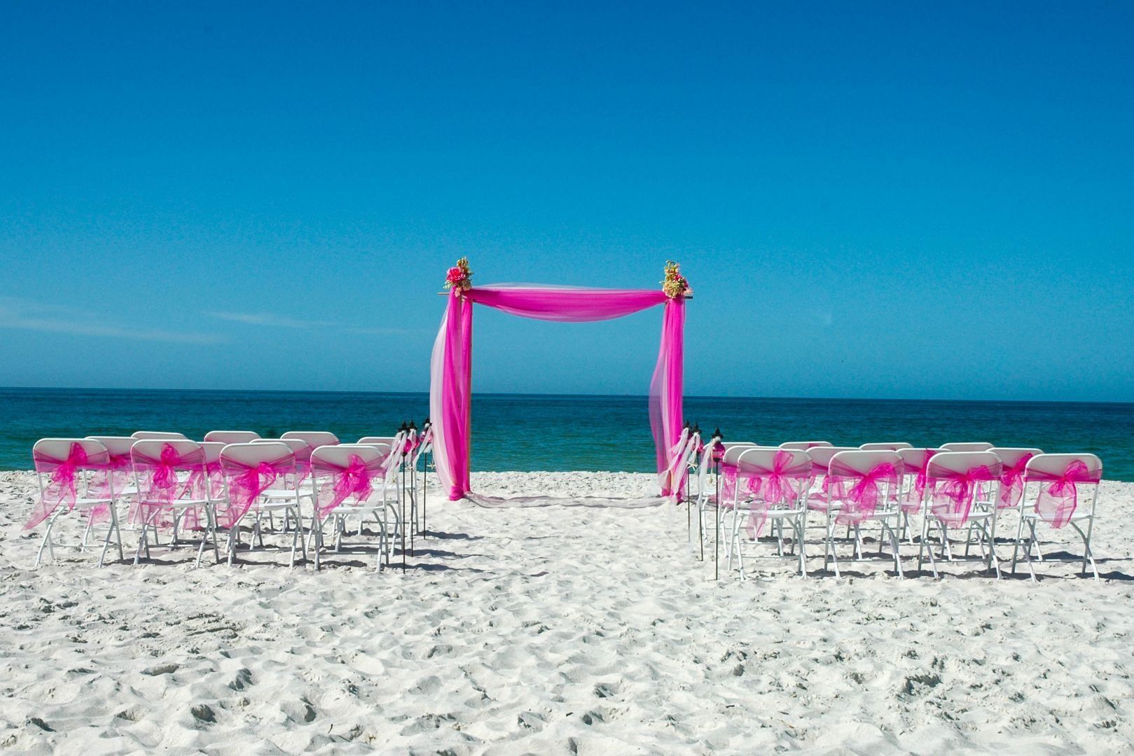 Beach wedding setup with pink decorations against a bright blue sky and ocean.