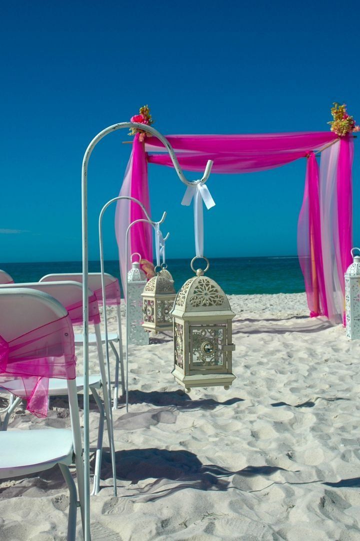 Beach wedding setup: white chairs, pink and white fabric draped arch, lanterns, blue sky, white sand.