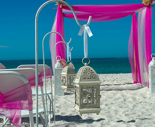 Beach wedding setup with pink draping, white chairs, and lanterns against a blue sky.