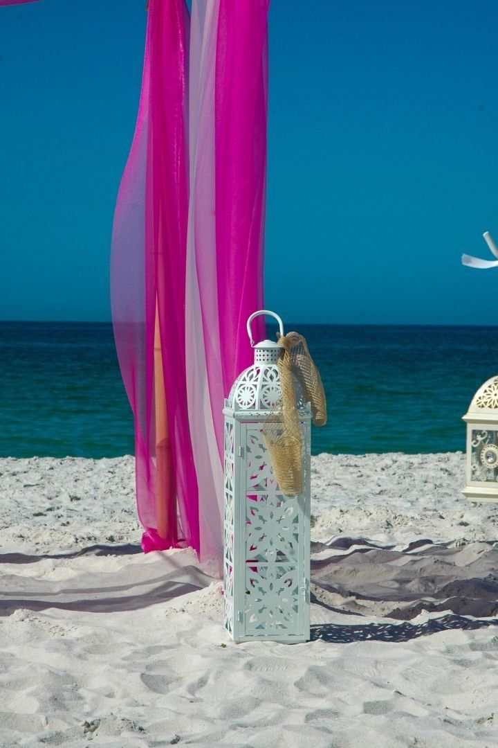 White lantern on beach with pink and white draped fabric, ocean, and blue sky background.