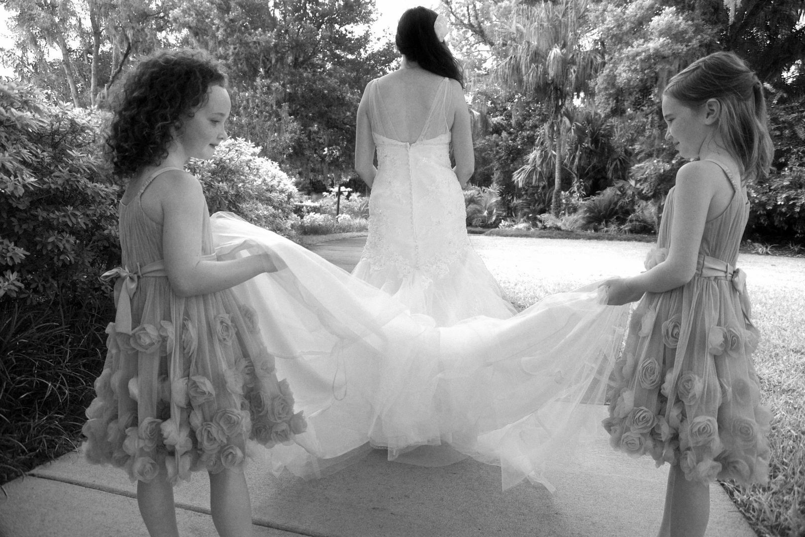 Bride in wedding dress, two flower girls holding her train outdoors in garden.