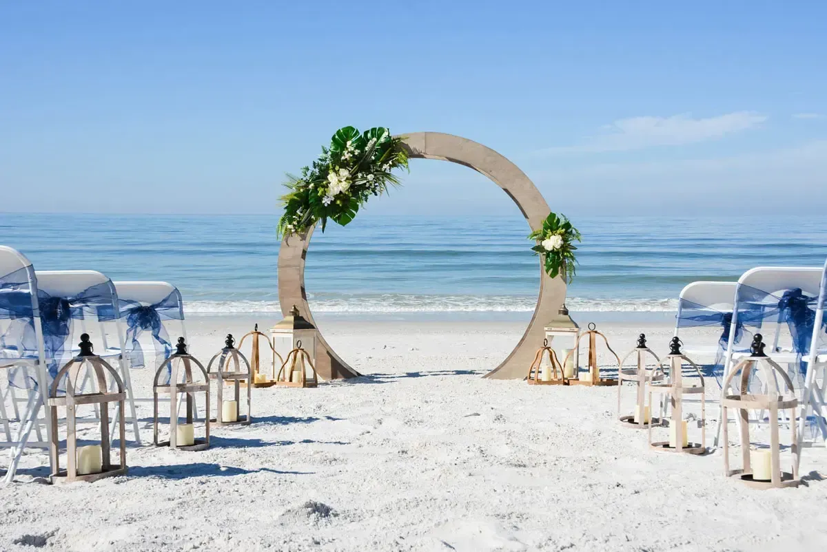 Beach wedding ceremony setup with a floral-decorated circular arch, chairs, and lanterns on the sand, with ocean in the background.