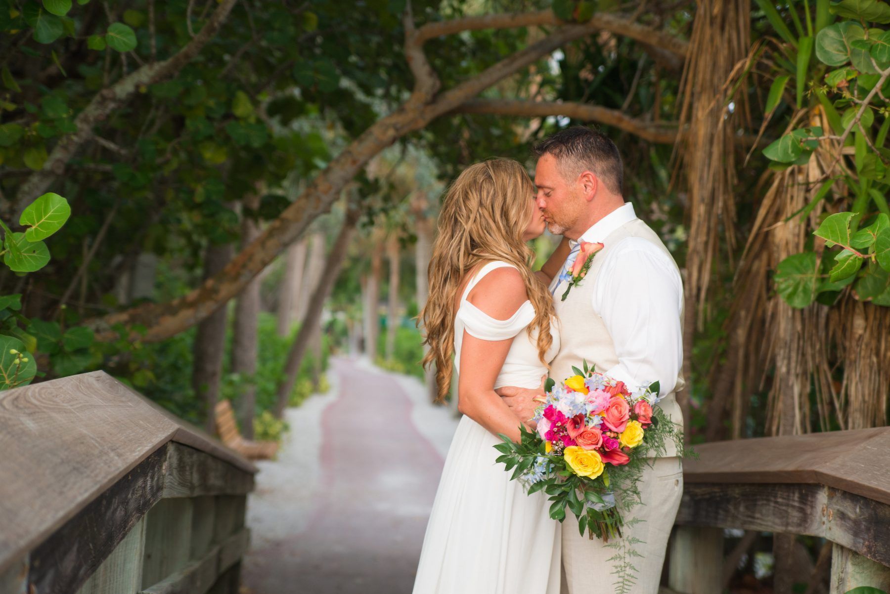 Couple kissing during their wedding, holding a bouquet, on a bridge surrounded by trees.