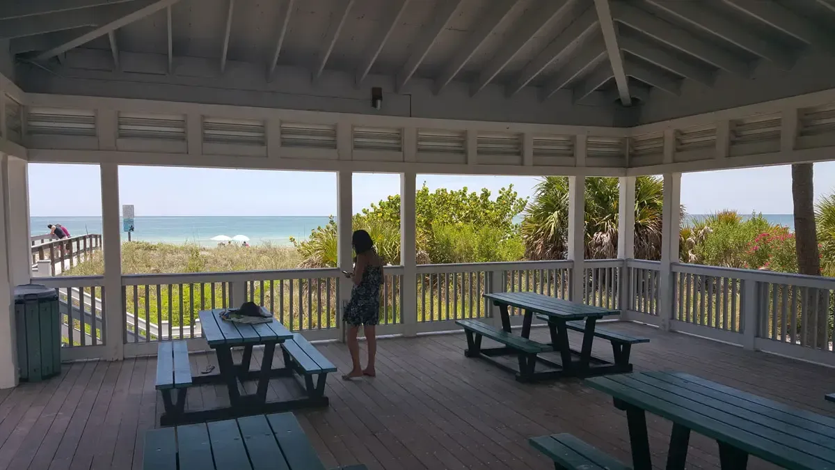 Covered picnic area overlooking the ocean. A person stands near a table; other tables are empty.