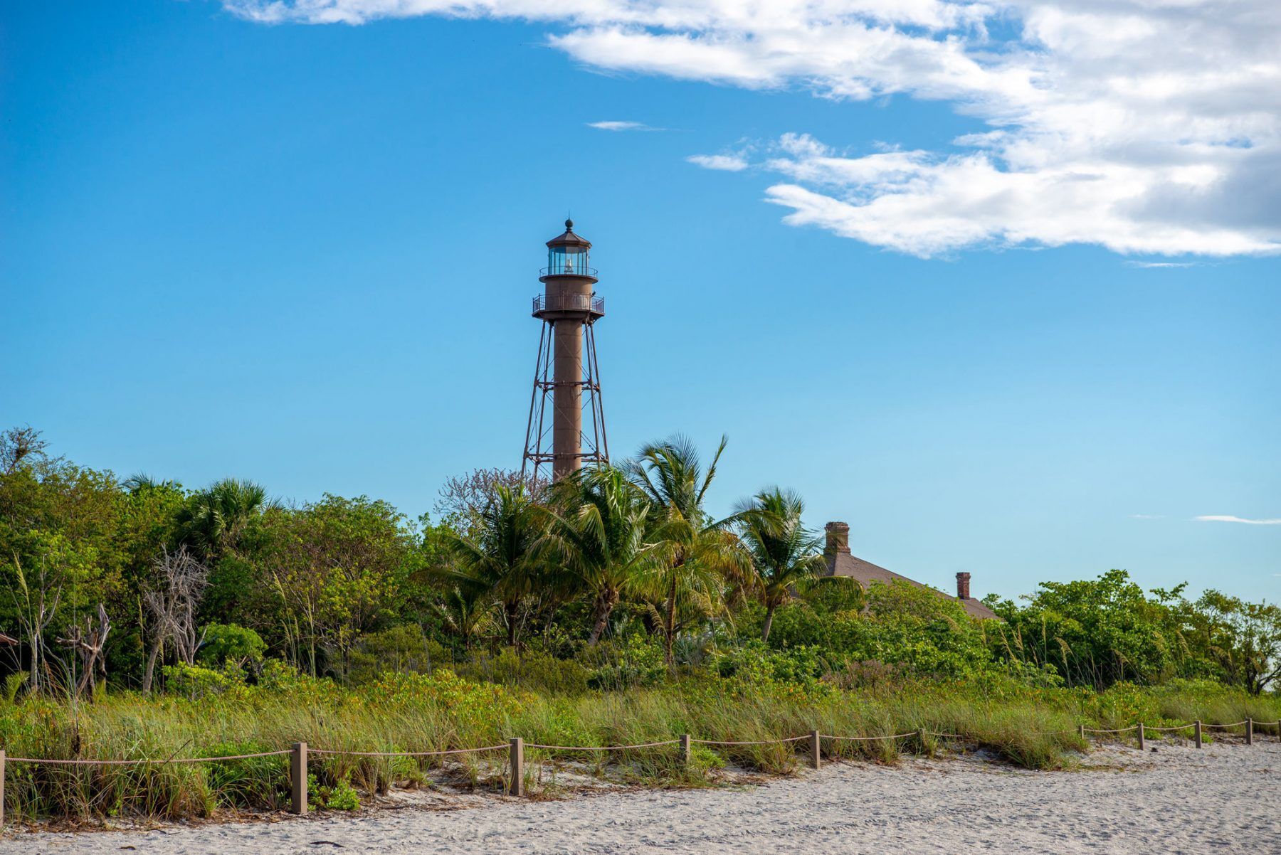 Lighthouse on a sandy beach under a blue sky, surrounded by green trees and foliage.