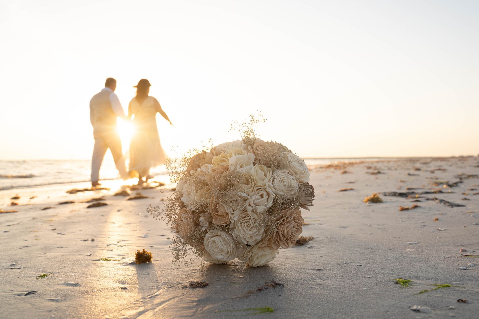 Wedding bouquet on beach, with silhouetted couple walking towards the sunset.