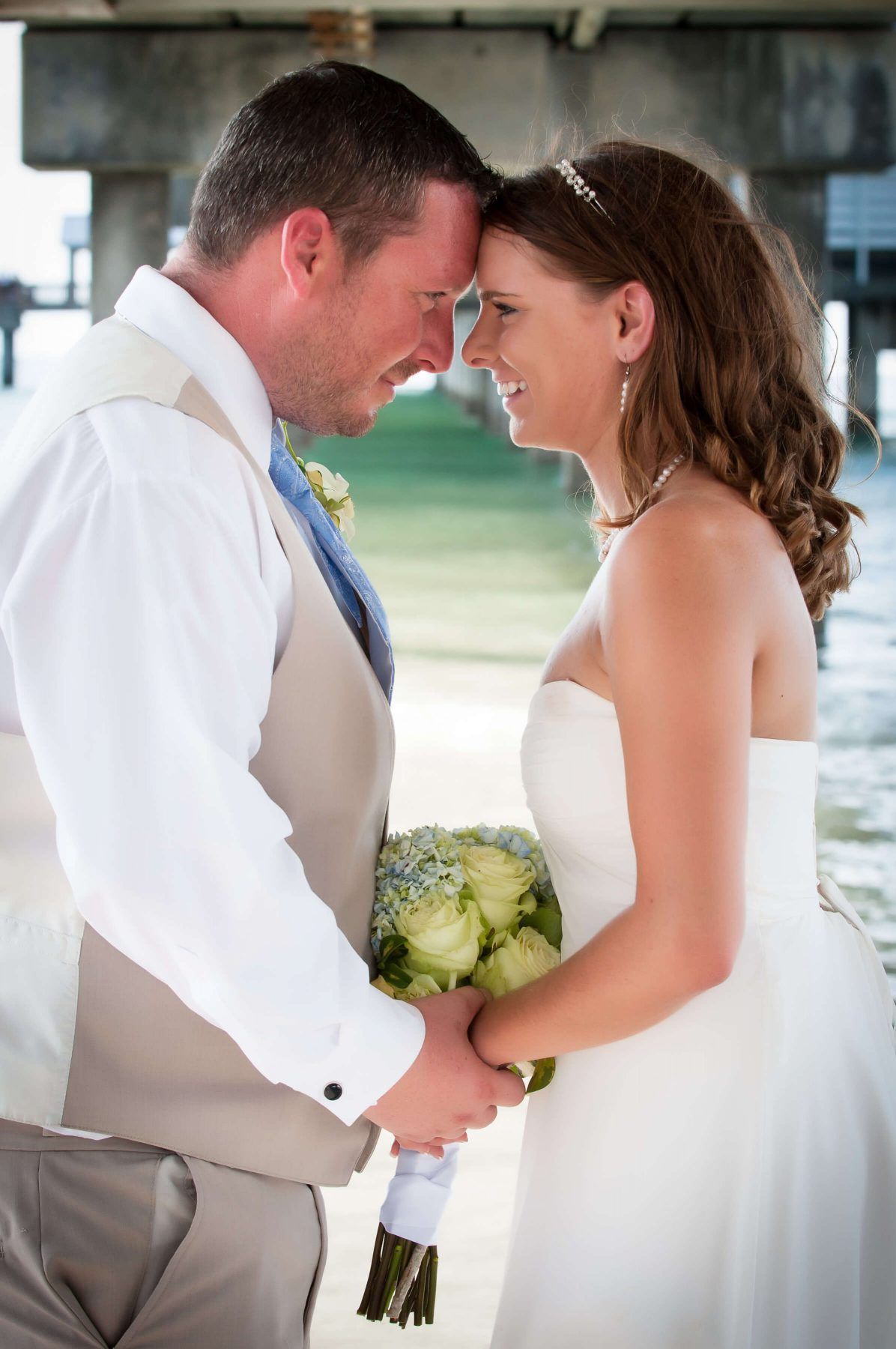 Bride and groom holding hands, foreheads touching, smiling, under a pier. Bride in white dress, groom in vest.