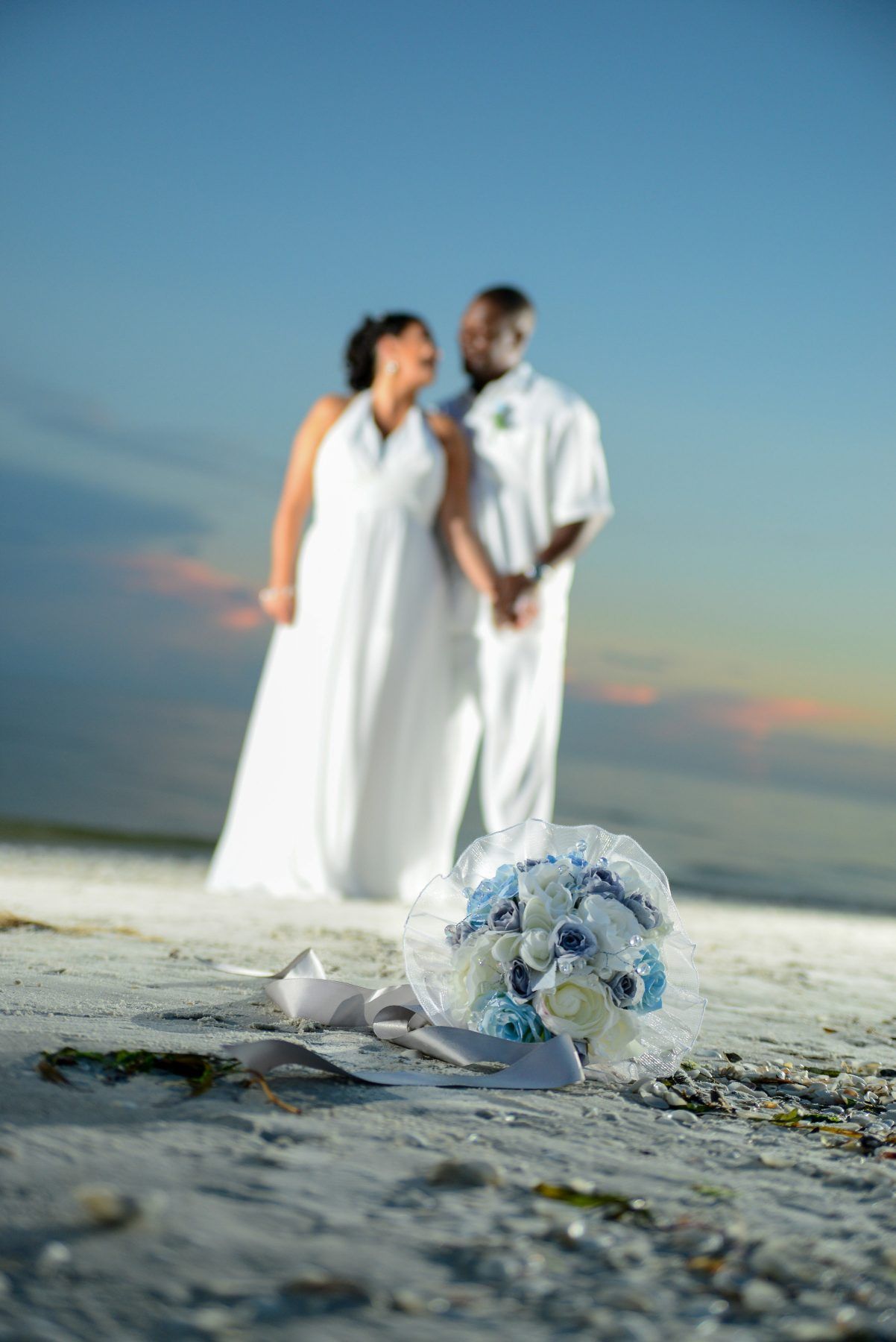 Wedding bouquet on beach, couple in background holding hands, sunset.